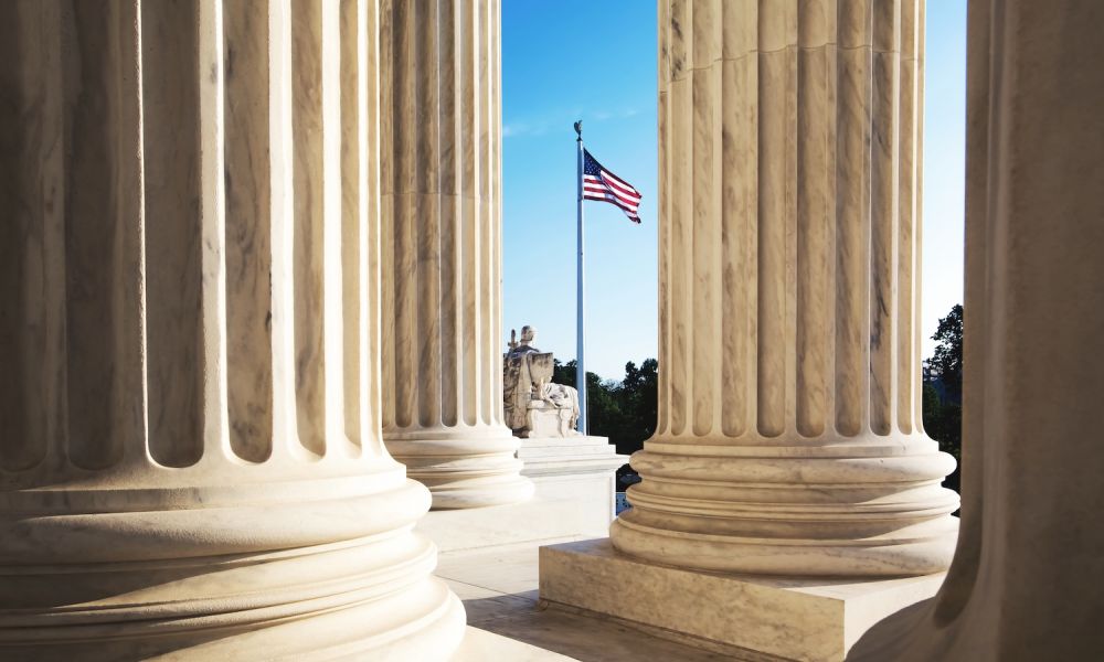 View through marble columns with US flag flying against a clear blue sky; statue visible between the pillars.
