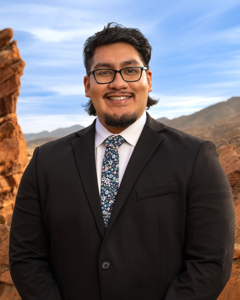 Man in a suit and floral tie smiles in front of scenic desert rocks and clear sky.