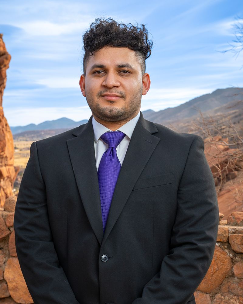 Man in suit with purple tie stands confidently against scenic rocky mountain backdrop.