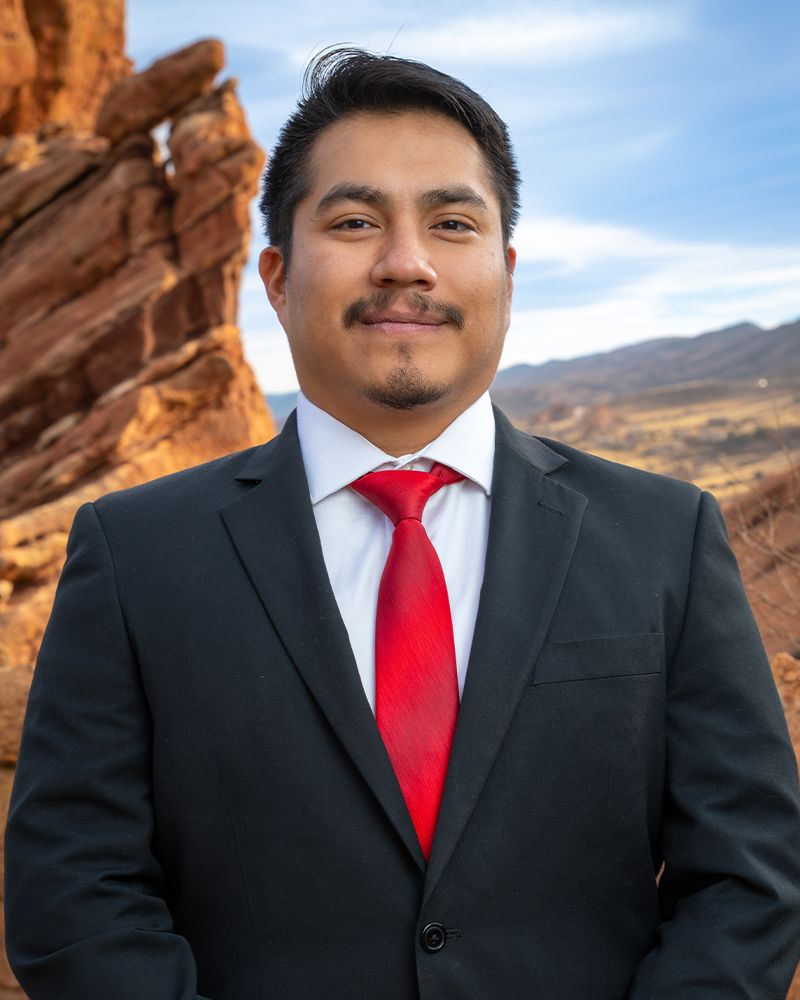 Man in a suit with a red tie standing in front of scenic, rocky landscape under a blue sky.