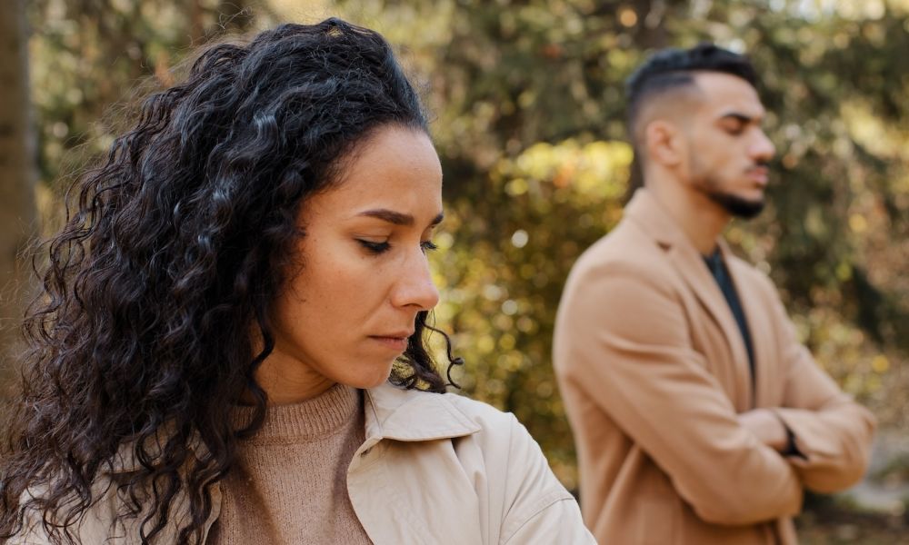 Two people standing with arms crossed, looking away from each other in a disagreement, surrounded by trees.