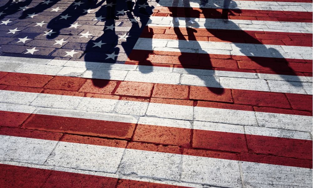 Shadows of people walking on an American flag painted on brick pavement, symbolizing unity and patriotism.