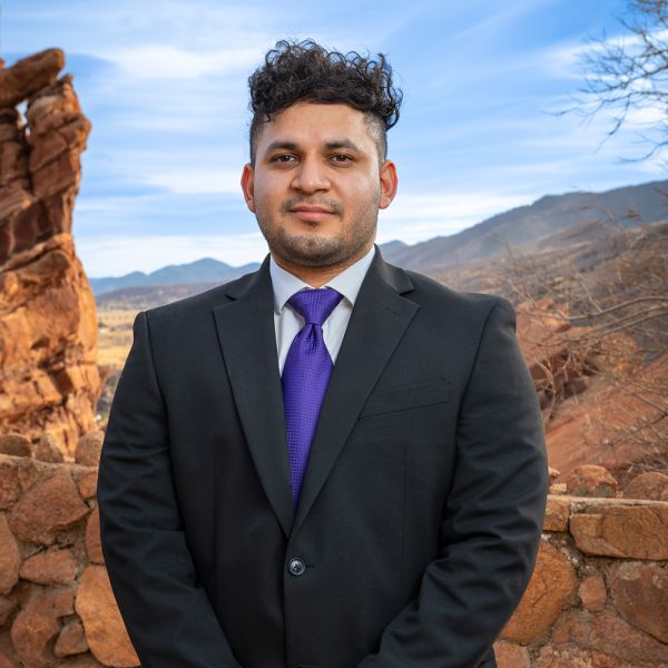 Man in suit with purple tie stands confidently against scenic rocky mountain backdrop.