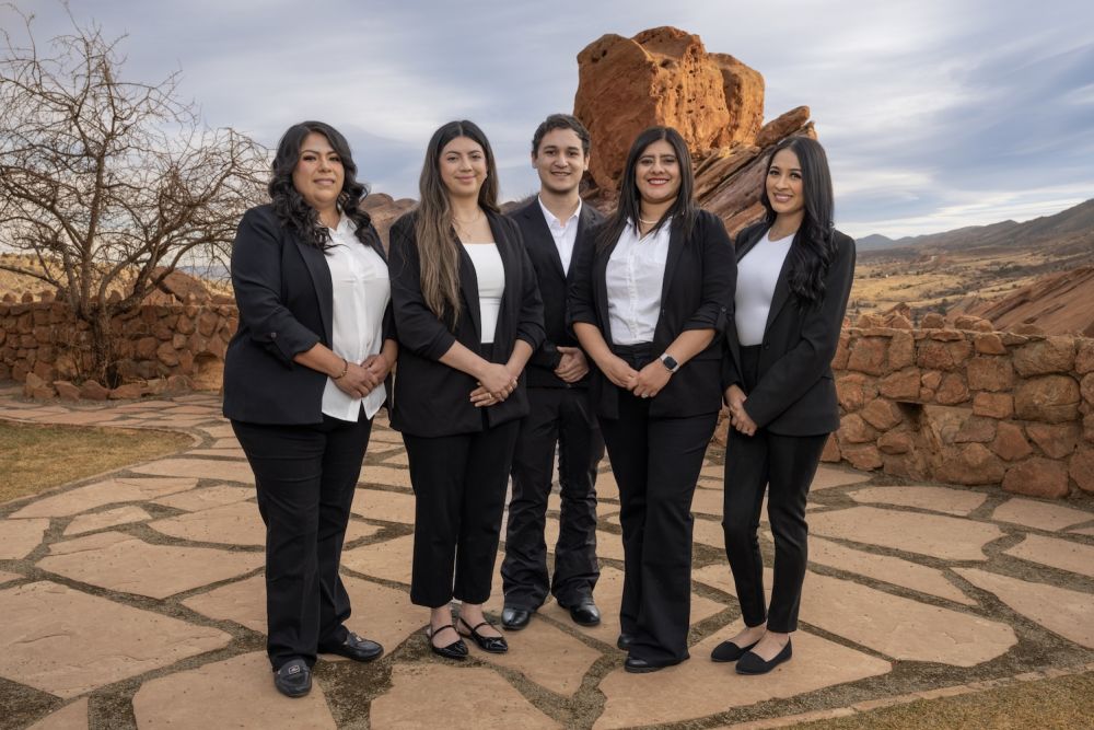 Five professionals in formal attire posing outdoors against a scenic rocky landscape.