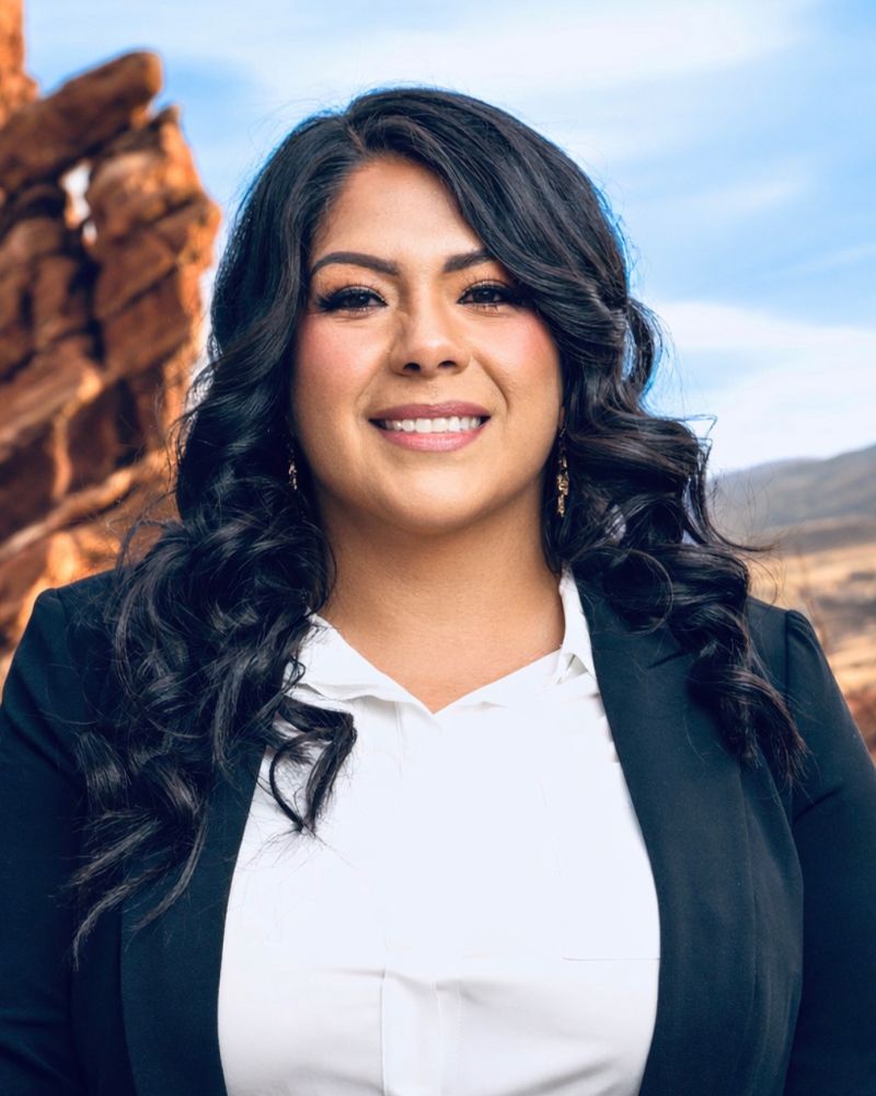 Smiling woman in business attire standing in a rocky, desert landscape under a clear blue sky.