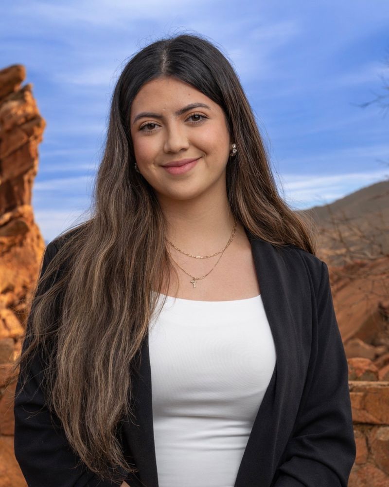 Woman smiling in front of red rock formations and blue sky, wearing a black blazer and white top.