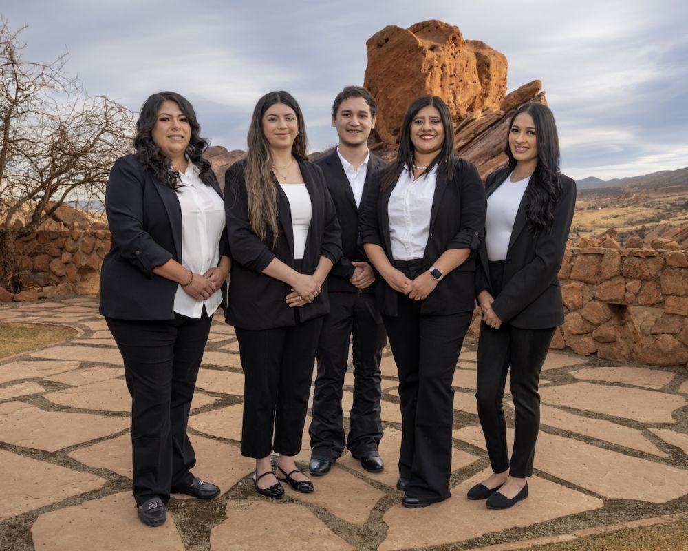 Five professionals in formal attire posing outdoors against a scenic rocky landscape.