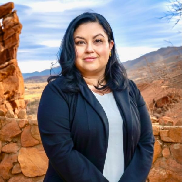 Woman in a black blazer standing outdoors with rocky landscape and blue sky in the background.