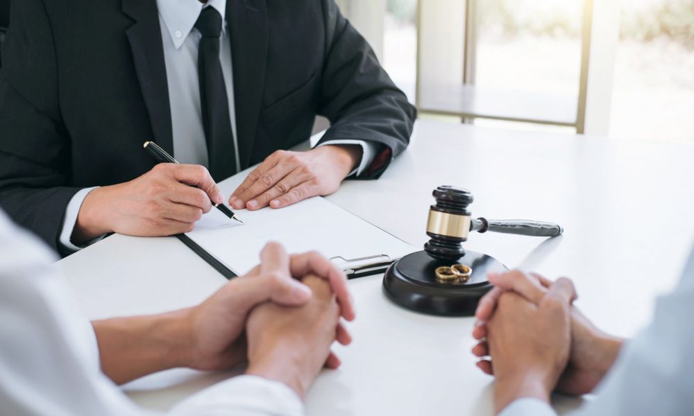 Lawyer signing divorce papers with clients, gavel and rings on table.