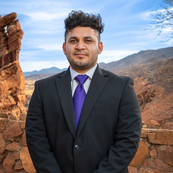 Man in suit with purple tie stands confidently against scenic rocky mountain backdrop.