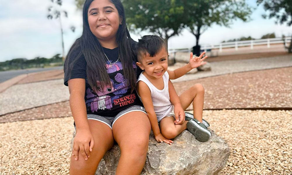 Kids sitting on a rock in a park, smiling at the camera with trees and a path in the background.