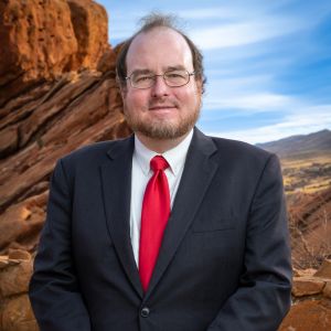 Man in a suit and red tie standing in a rocky outdoor setting under a blue sky.