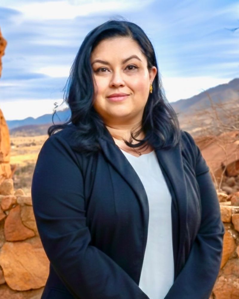 Mujer con saco negro al aire libre con un paisaje rocoso y un cielo azul al fondo.