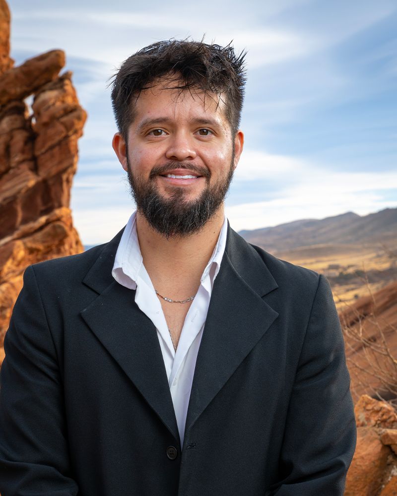 Man in a suit smiling in front of scenic rocky landscape and blue sky.