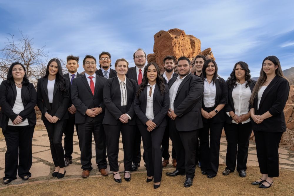 A professional team stands outdoors, dressed in formal attire, with a rocky landscape backdrop and a clear sky.