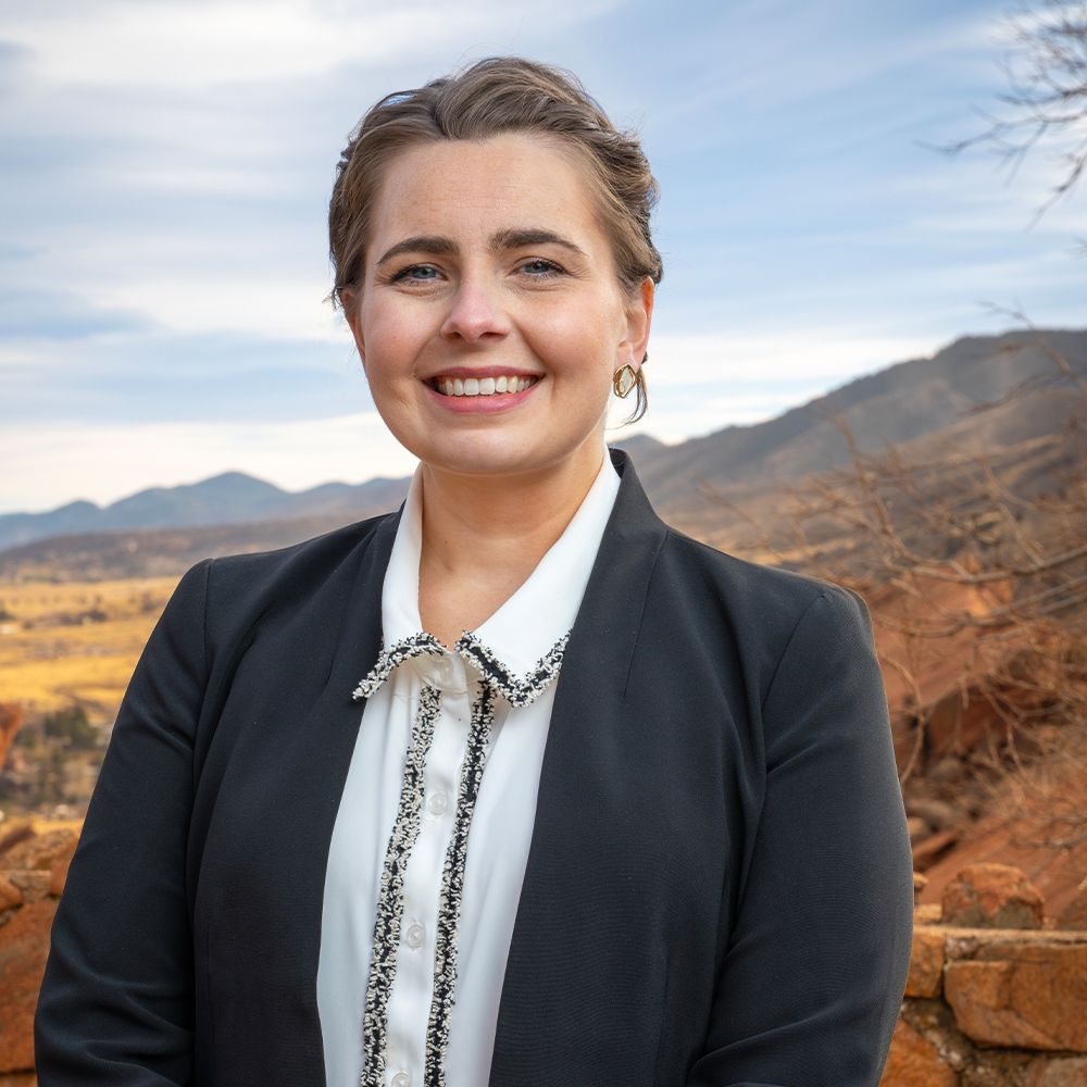 Smiling person in a black blazer with a scenic mountain landscape background.