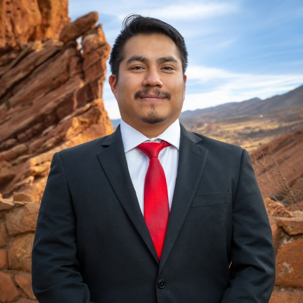Man in a suit with a red tie standing in front of scenic, rocky landscape under a blue sky.