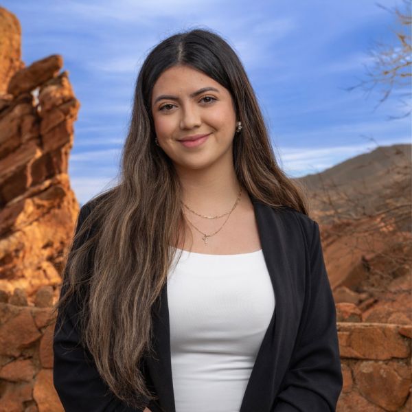 Woman smiling in front of red rock formations and blue sky, wearing a black blazer and white top.