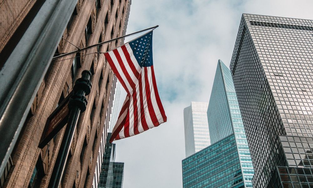 USA flag waving amidst tall urban skyscrapers against a cloudy sky backdrop, symbolizing American cityscape.