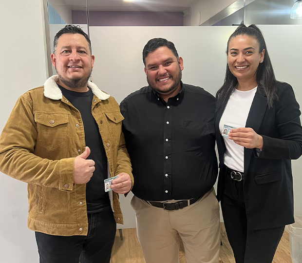 Two people holding ID cards and smiling beside a man in a black shirt in an office with frosted glass panels.