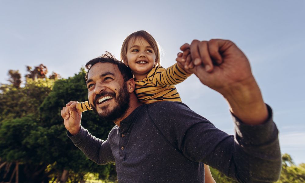 Father and child smiling outdoors, enjoying a sunny day with greenery in the background.