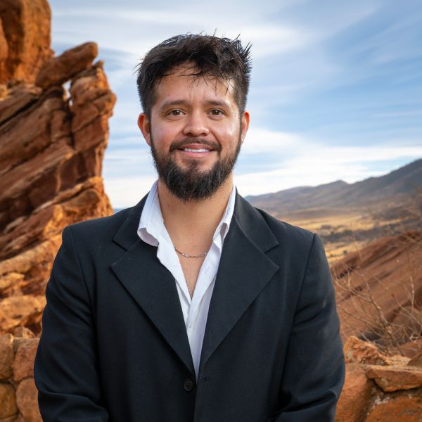 Man in a suit smiling in front of scenic rocky landscape and blue sky.