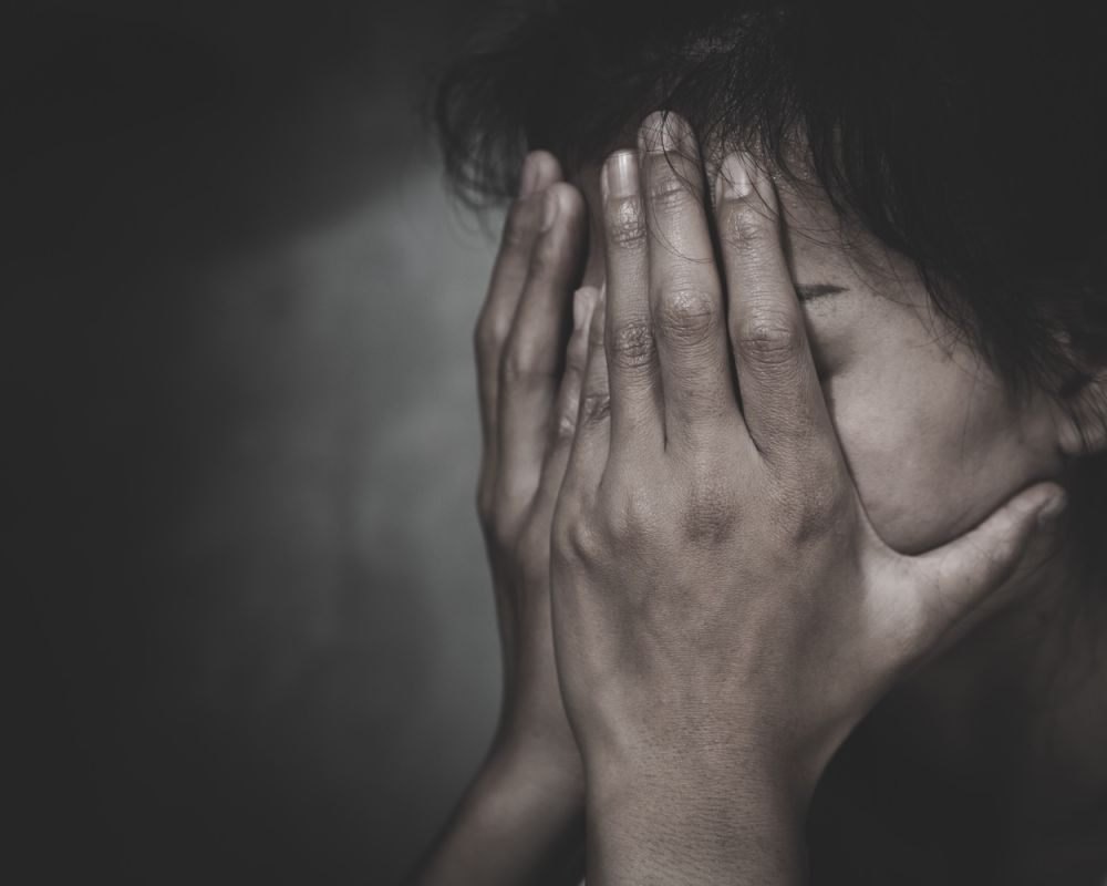 Person covering face with hands, expressing stress or anxiety in a dark, moody setting.