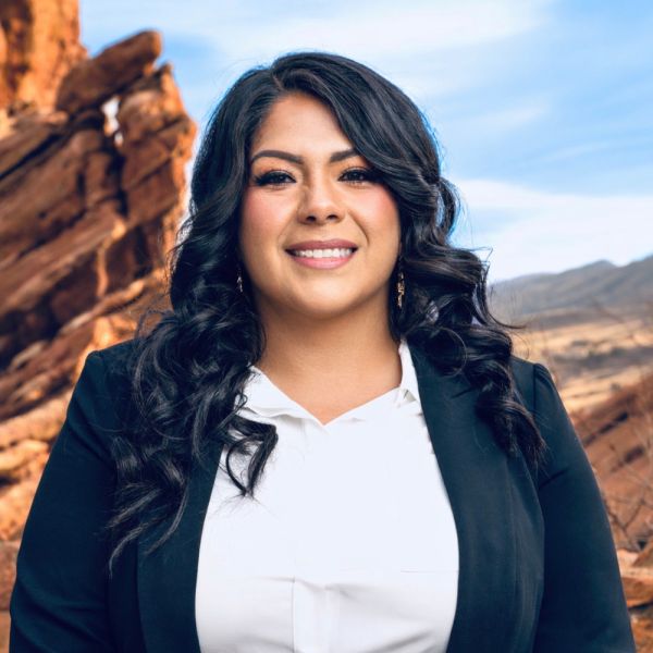 Smiling woman in business attire standing in a rocky, desert landscape under a clear blue sky.