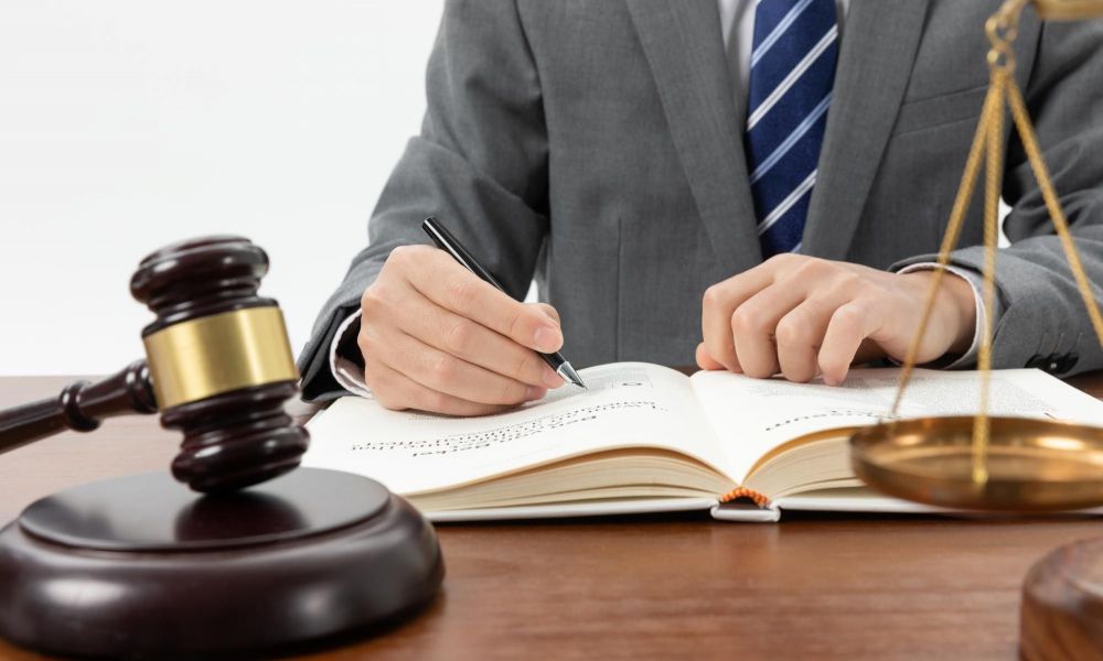 Lawyer writing in a book at a desk with gavel and scales of justice in foreground. Legal consultation scene.