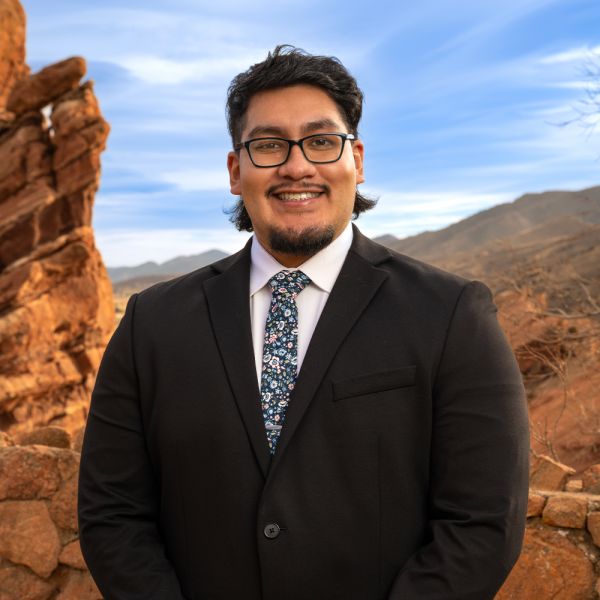 Man in a suit and floral tie smiles in front of scenic desert rocks and clear sky.