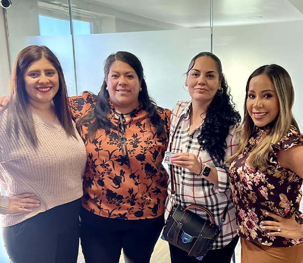 Four women standing together and smiling in an office setting, with one woman holding a small card.